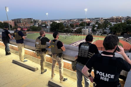 A line of officers in bulletproof vests stand on a balcony looking down at a football field.