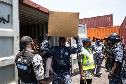 A man carries a box on his head, while armed police stand near him.