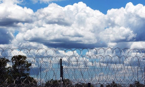 Razor wire fence at a prison, with treetops beyond it and a blue sky with large fluffy white clouds
