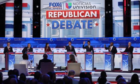 A broad, bright stage, mostly white with some red and blue, and a line of seven lecterns with an adult standing behind each one. Multiple people are raising their hands and speaking at the same time.