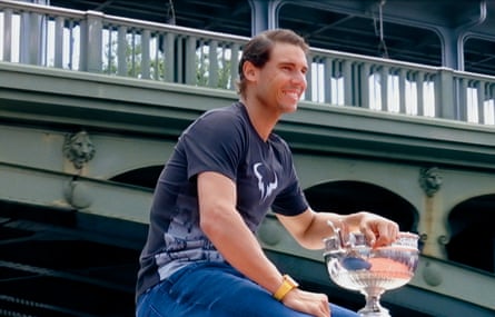 A man in a black T shirt and blue jeans holding a silver trophy cup with a bridge in the background