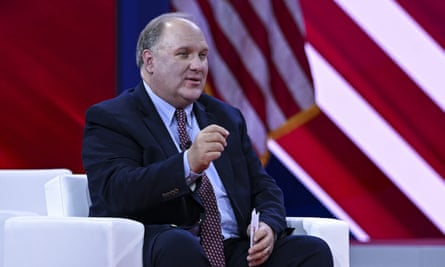 John Solomon speaking while seated on a stage with the US flag behind