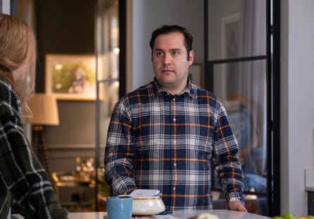 Michael Theo wearing a plaid shirt, looking serious, standing at a kitchen bar as if talking to someone.