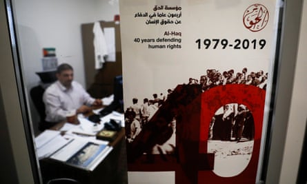 Photo of a glass door with a poster on it reading: ‘Al Haq: 40 years of defending human rights” in English and Arabic. Behind the glass door and out of focus, a man sits at a desk covered in papers