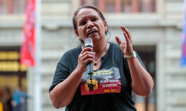 Megan Krakouer is speaking at an outdoor event, holding a cordless microphone in one hand and gesturing with the other