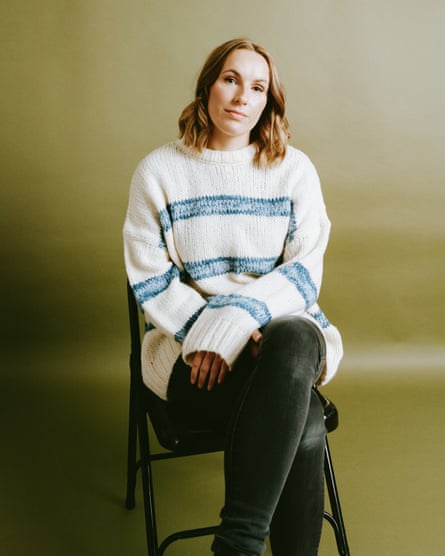 Rebecca Coxon sitting on a black chair against an olive-green background, wearing a baggy, white jumper with blue stripes and tight, black trousers, her hands placed in a relaxed manner on her lap