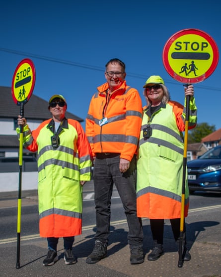 The two women, in their lollipop gear, holding sticks, stand either side of Patmore by a street crossing. All three are smiling at the camera
