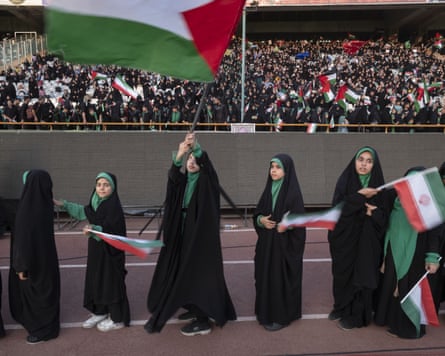 A group of girls wearing burqas waving Iranian and Palestinian flags in front of a large crowd