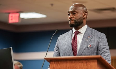 A Black man with a beard wearing a grey suit and red tie stands with lips pressed together at a microphone at a desktop lectern.