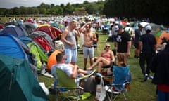 Tennis fans camp in the queue: lines of tents are seen in a park opposite the tennis club grounds; people are relaxing in camping chairs and standing talking to one another and look happy and sociable. It is sunny and most are wearing shorts and T-shirts, some with baseball caps and sunglasses. One young man is barechested in denim shorts.