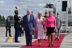 Charles, Camilla and Élisabeth Borne walk along a red carpet on the airport tarmac, with the plane and soldiers in ceremonial uniform in the background