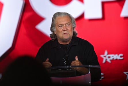 Steve Bannon speaks at a podium, gesturing with both thumbs up, against a red CPAC backdrop