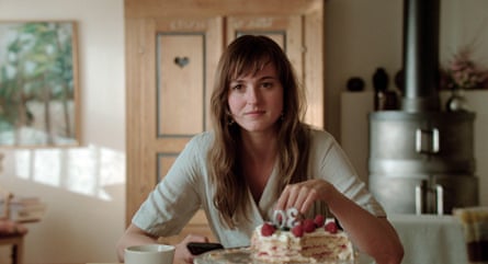 she sits at a kitchen table in front of a stove and wooden door, with a slice of birthday cake in front of her with the number 30 on it. She has long light brown hair with a fringe and wears a pale green wrap-style top