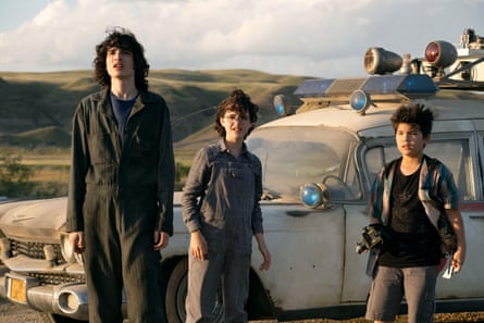 A tall man with dark curly hair, standing next to two children in front of an old white car, with a vast landscape behind them