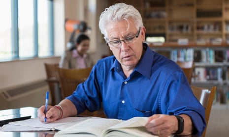 Older man reading book in library and writing notes