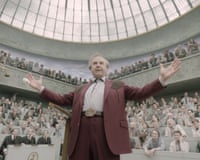 Tom Cruise as Digger Rockwell raises both arms while speaking in a domed, crowded auditorium
