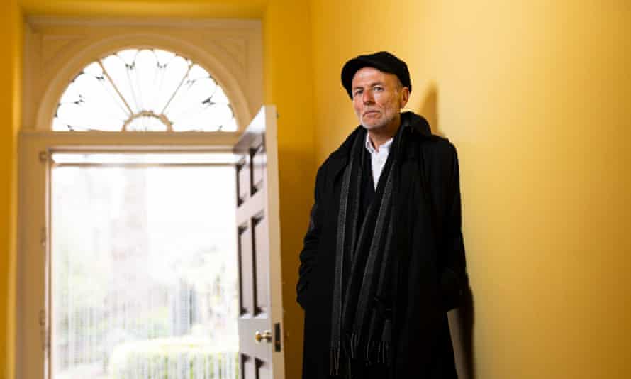 Graham Caveney in a long dark coat, scarf and hat, standing in a yellow pained hallway near a door, open to the outside