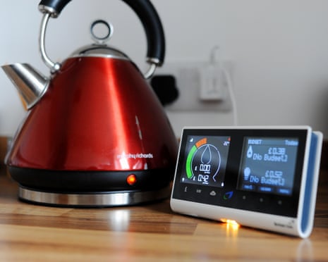 A red kettle sits beside a digital smart meter display showing energy usage on a kitchen counter