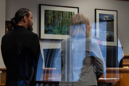 King County Sheriff’s Deputy Ricardo Arturo Cueva, a man with light-toned skin, a beard and hair in a ponytail, wears a suit, and stands facing away from the camera. To his right stands a woman with light-toned skin and gray, medium-length hair, wearing a gray suit jacket.