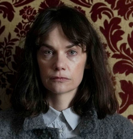 Ruth Wilson, looking unhappy, in a cardigan and buttoned-up shirt against flocked wallpaper