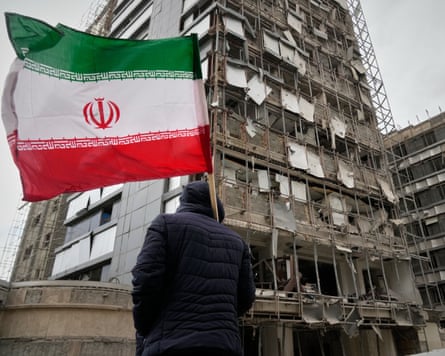 Man holding Iranian flag in front of damaged hospital facade.