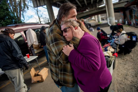 A person with long brown hair who is wearing a flannel shirt hugs another person who appears grief-stricken and wearing sunglasses