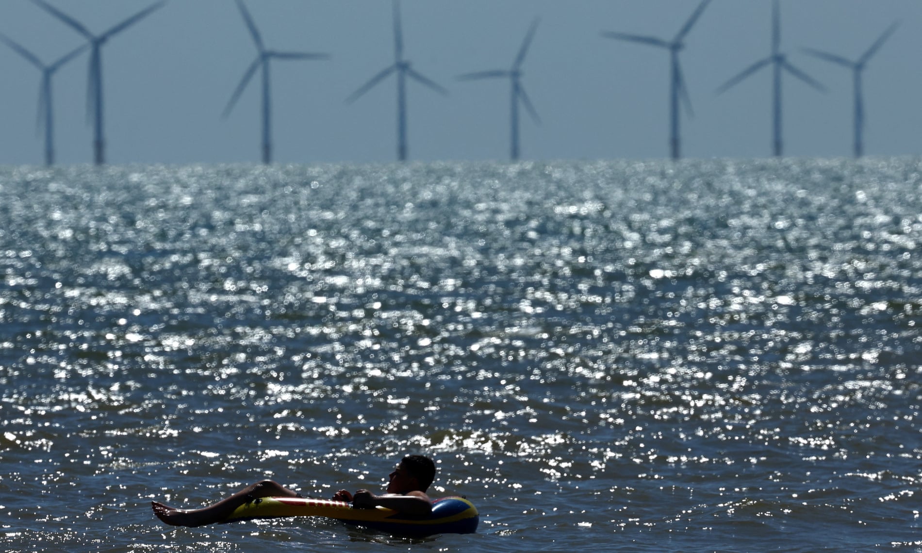 Un hombre flota en un inflable sobre el agua en un día soleado, mientras en el horizonte se observan varias turbinas eólicas marinas girando en el mar. El agua brilla intensamente por los reflejos del sol, creando un contraste con las siluetas de las turbinas en el fondo.