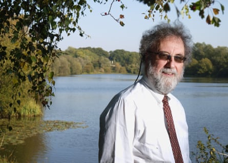 Robert Watson stands in front of a lake with trees on the far side, an autumnal light.