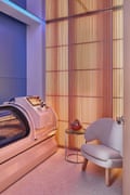 A hyperbaric chamber in a softly lit room, with a table and off-white cushioned chair next to it