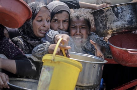 Palestinians including children jostle together as they hold out containers for food