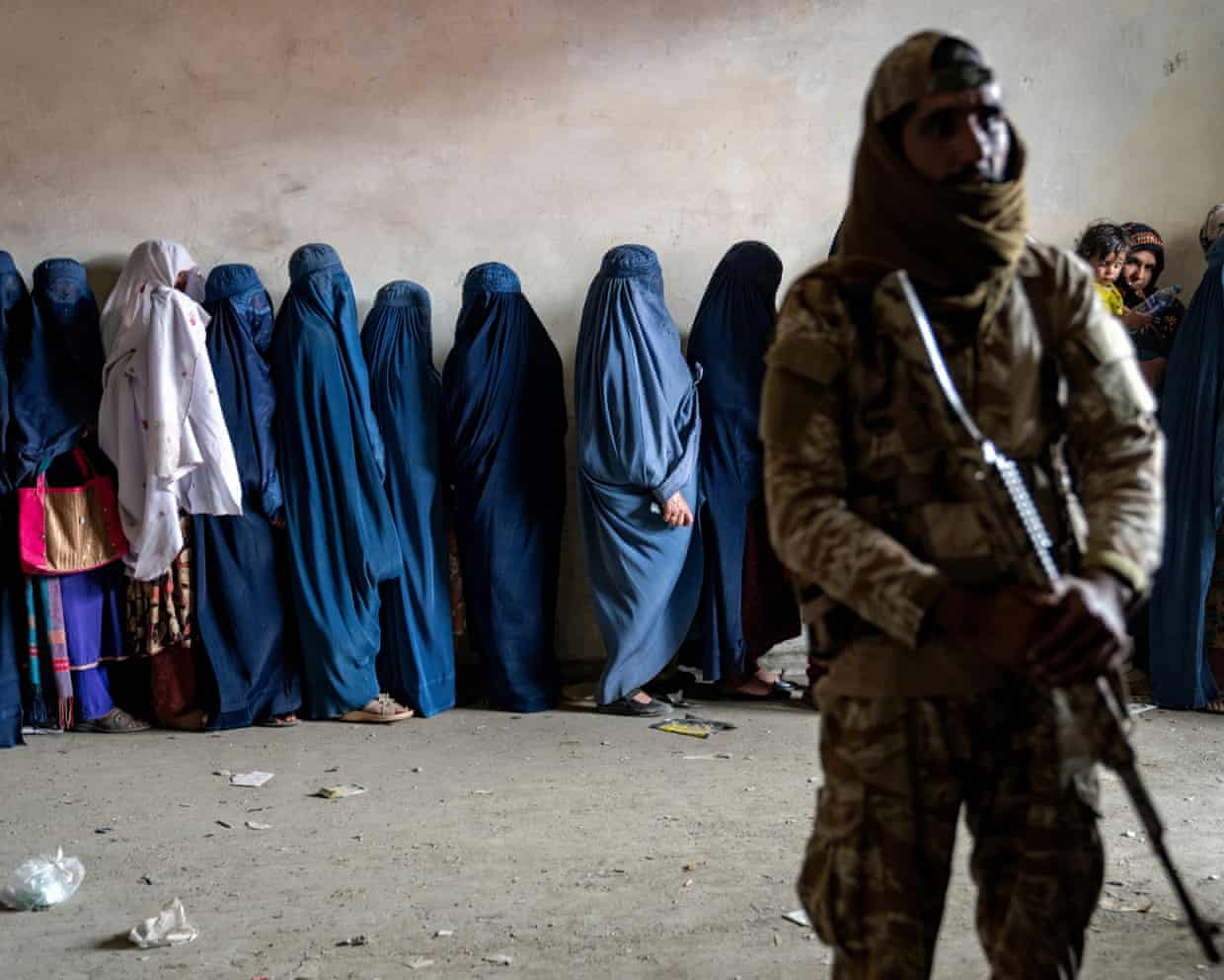 Afghan women wearing burqas stand in a line beside a soldier in uniform holding a rifle.