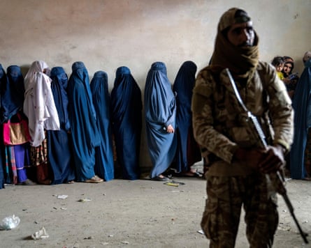 A Taliban fighter stands with a machine gun while women whose bodies are entirely covered wait in a queue
