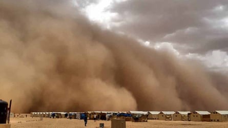 A line of tin huts on sand with a huge cloud of billowing dust about to engulf them