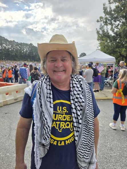 man with cowboy hat, a ‘US murder patrol’ shirt and keffiyeh around his shoulders smiles at protest