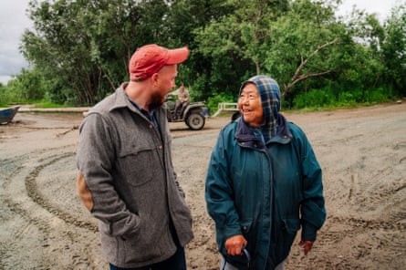 A man in a coat and baseball hat walks and smiles with a woman with her head covered in a scarf, also smiling, across muddy ground.