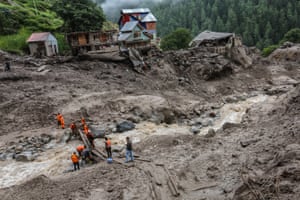 Debris is strewn around muddy banks of a fast moving stream as rescuers work at the flood site with the remains of building in the background