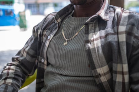 A man, whose face cannot be seen, wearing a plaid shirt over a grey tshirt with a gold chain around his neck, bearing a letter H.