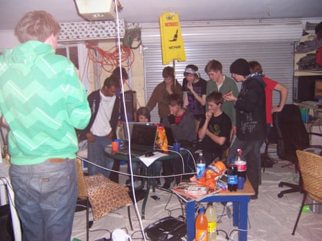 A group of young people, mostly boys, gathered round some computers in a space that looks like a garage, with cables everywhere and fizzy drinks and snacks on a table