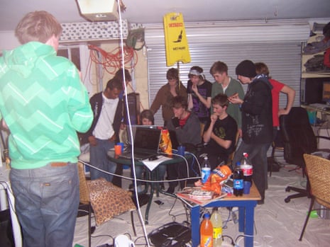 A group of young people, mostly boys, gathered round some computers in a space that looks like a garage, with cables everywhere and fizzy drinks and snacks on a table