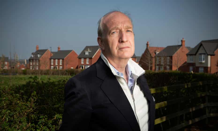 Portrait of Nigel Hugill in a jacket and open-necked shirt in the foreground, looking at the camera, with new-build  houses visible in the background