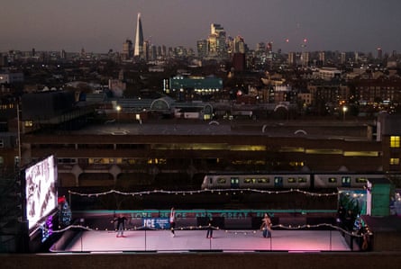 An aerial photo at night of the London skyline, with skyscrapers in the distance and a small ice rink with a big screen at one end on a roof in the foreground