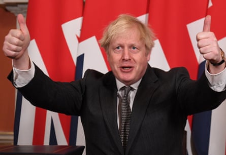 A man in a dark suit and tie, with blond hair, holding both thumbs up. A union jack flag is behind him