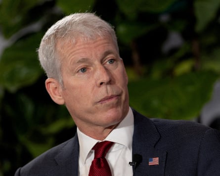 Chris Wright is wearing a suit and has a small badge of the US flag pinned to his lapel