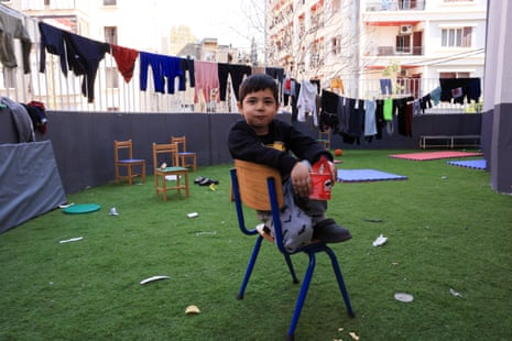 A small boy sits with his legs curled up on a chair in the grass of a courtyard. Behind him are more chairs, some mats, a mattress and a clothesline that is heavy with colourful clothes.