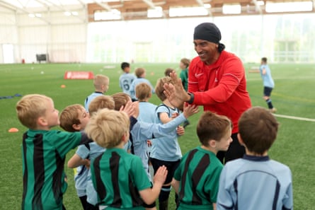 A youth coach, wearing a red training top, high fives members of two 3-a-side teams made up of young children. One team is wearing green shirts with black vertical stripes, the other light blue shirts.