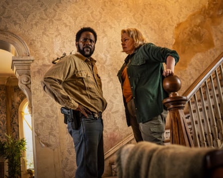 A still from a show called From showing a black man in jeans and a jacket speaking to an older white woman who is standing on the bottom step of a staircase