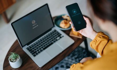 Young Asian woman logging in to her laptop and holding smartphone on hand with a security key lock icon on the screen, sitting in the living room at cozy home. Privacy protection, internet and mobile security concept