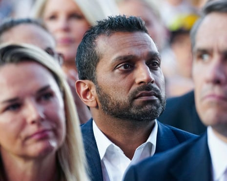 Kash Patel, wearing a navy suit and a white shirt, waits in a crowd of people.