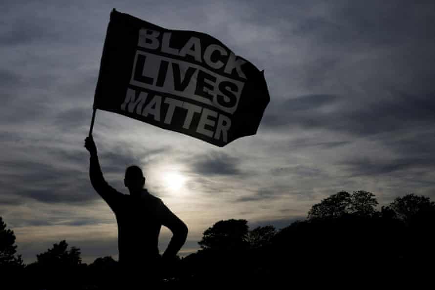 Silhouette of a man waving a large Black Lives Matter flag against a background of the setting sun.