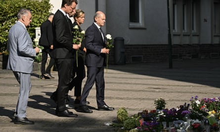 Olaf Scholz stands holding a white rose with several other people in front of floral tributes laid out on the floor